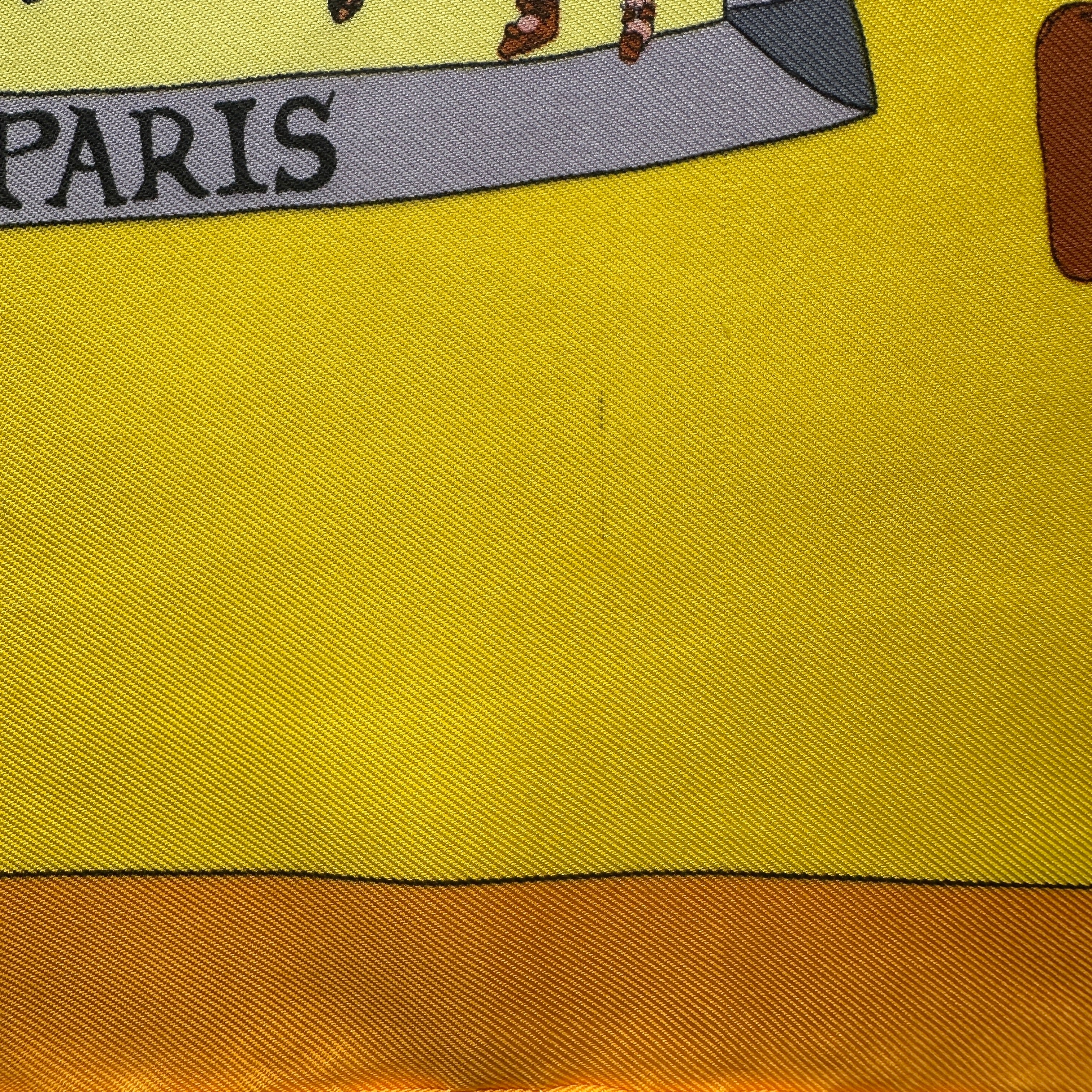 Détail d'une minuscule marque ou point sombre isolé sur le fond jaune, situé au-dessus de la signature Hermès Paris.