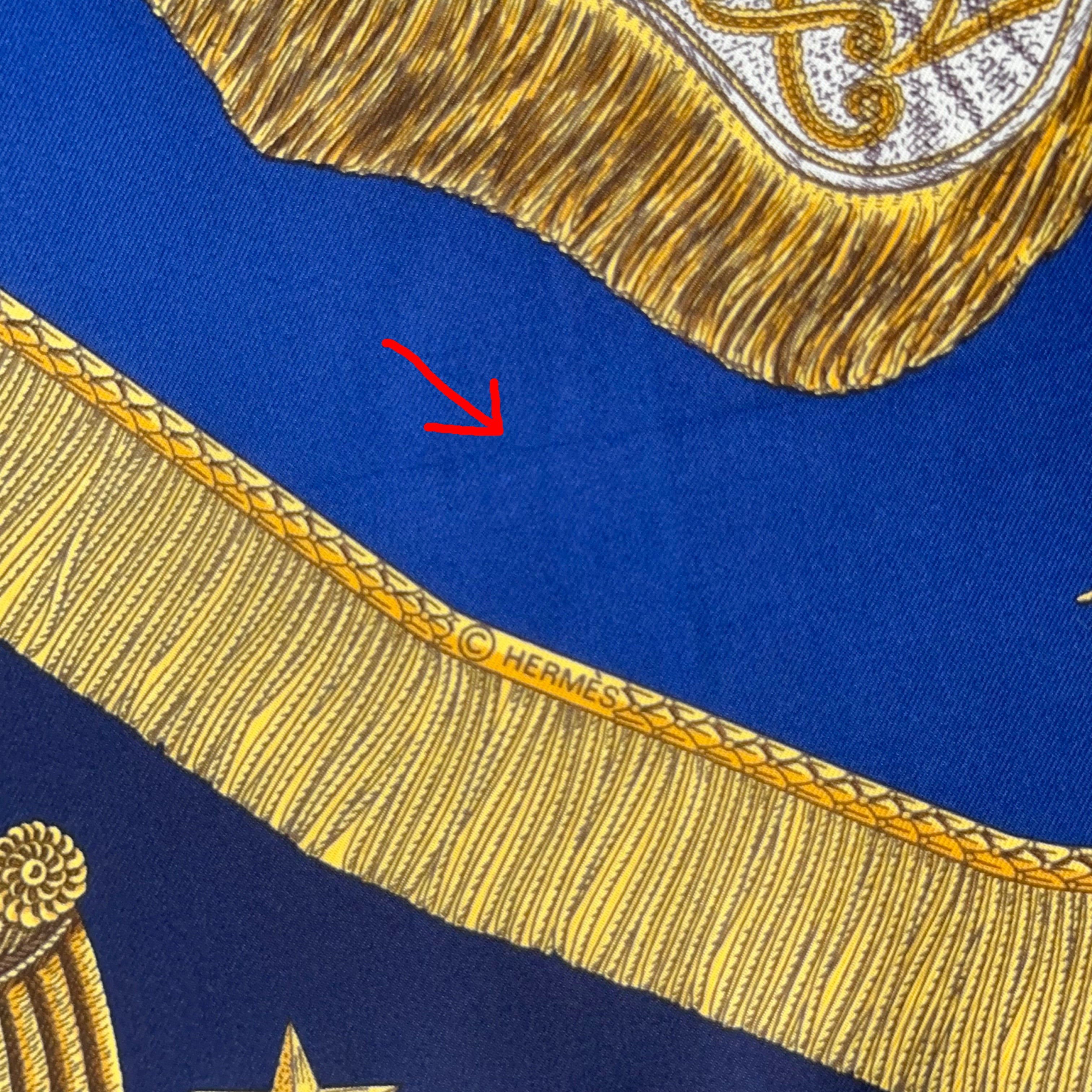 Zoom macro sur la bordure à franges dorées en trompe-l'œil, incluant le copyright "© HERMÈS" discret.