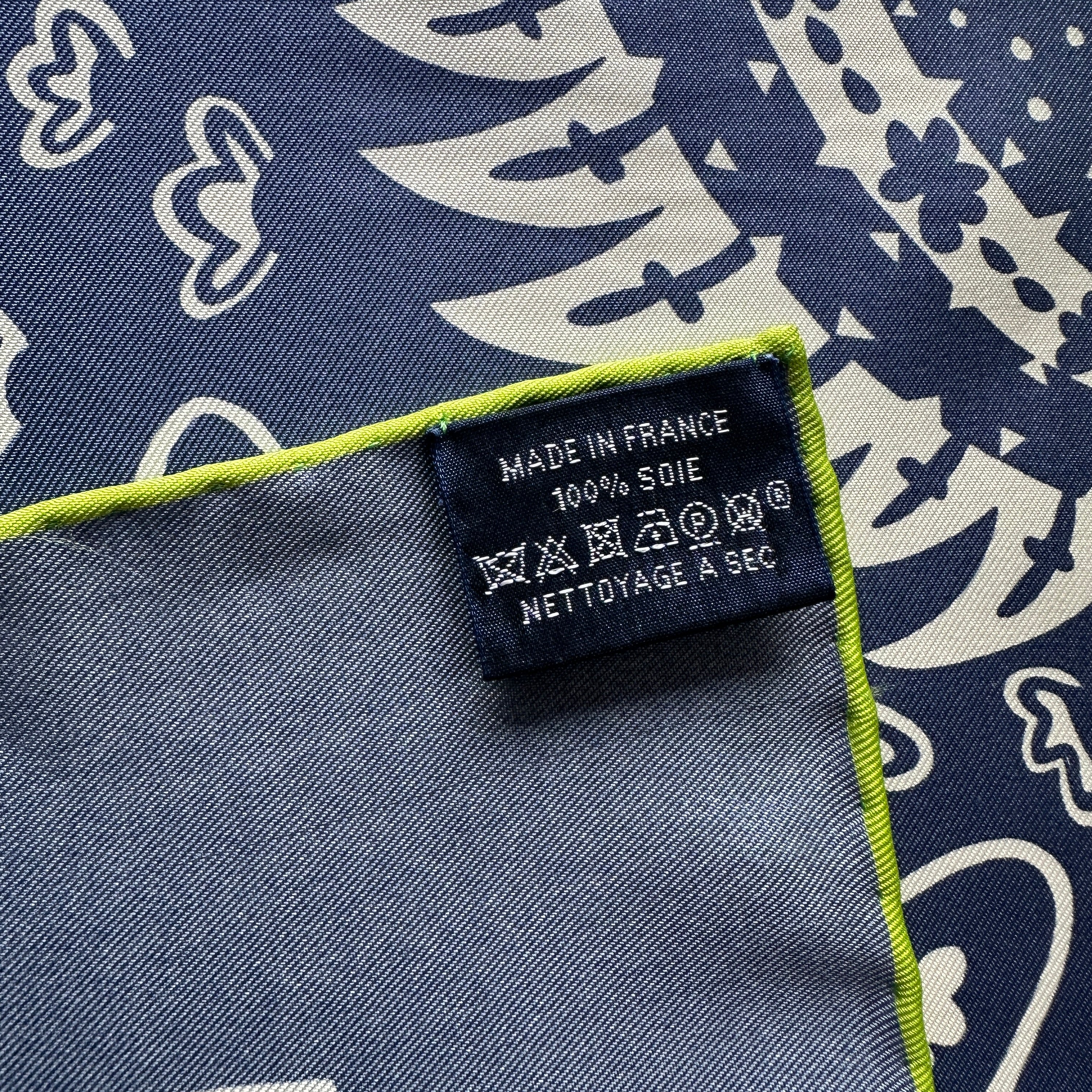 Étiquette d'entretien noire cousue sur le bord vert du foulard Hermès, indiquant "MADE IN FRANCE", "100% SOIE" et les symboles de nettoyage à sec.